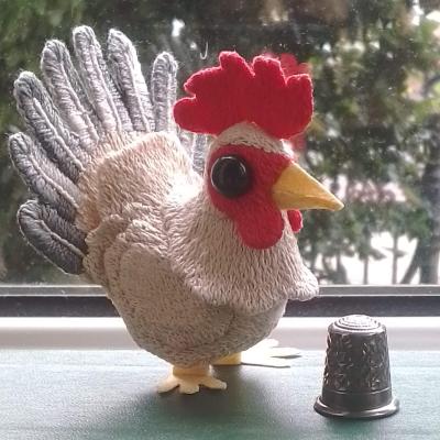 A white cockerel with grey tail feathers and red crest stands beside a thimble (which is about a third of its height)